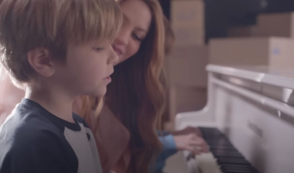 Delante de un piano tocando. Primer plano de Sasha Piqué Mebarak en el videoclip de Acróstico. En segundo plano aparece Shakira observándole con cariño. Al fondo las manos de Milan Piqué tocando el piano. 